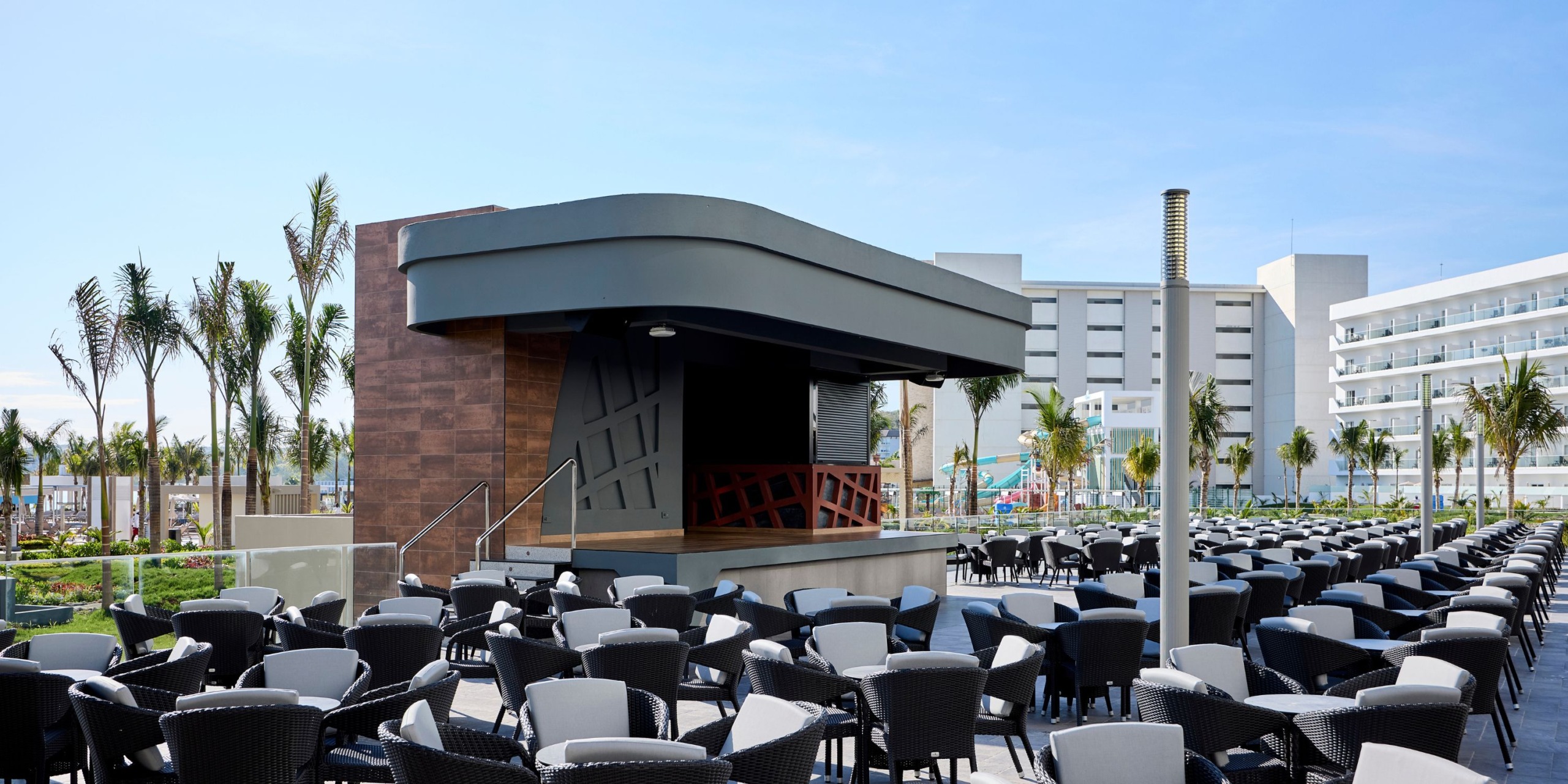 The outdoor seating area at Hotel RIU Palace Aquarelle features rows of black tables and chairs facing a closed bar, with palm trees and modern buildings gracing the background under a clear blue sky.