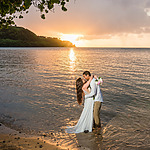 A couple stands embraced in shallow ocean water during sunset, the sun setting behind a hilly coastline. The bride holds a bouquet of flowers, both dressed in wedding attire, capturing the essence of destination wedding sunsets.