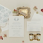 Flat lay of wedding stationery, including an invitation, RSVP card, decorative envelope, gold tray, ribbon, and vintage binoculars surrounded by dried flowers on a beige background.