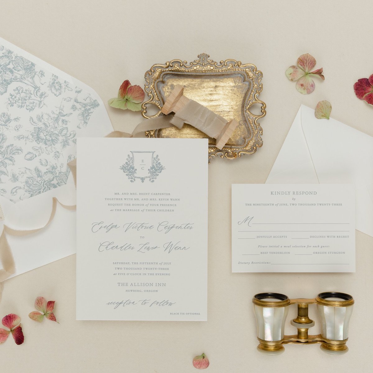 Flat lay of wedding stationery, including an invitation, RSVP card, decorative envelope, gold tray, ribbon, and vintage binoculars surrounded by dried flowers on a beige background.