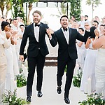 Two grooms in black tuxedos walk down the aisle hand-in-hand, surrounded by applauding guests in a stunning tropical outdoor setting, reminiscent of glamorous celebrity weddings.