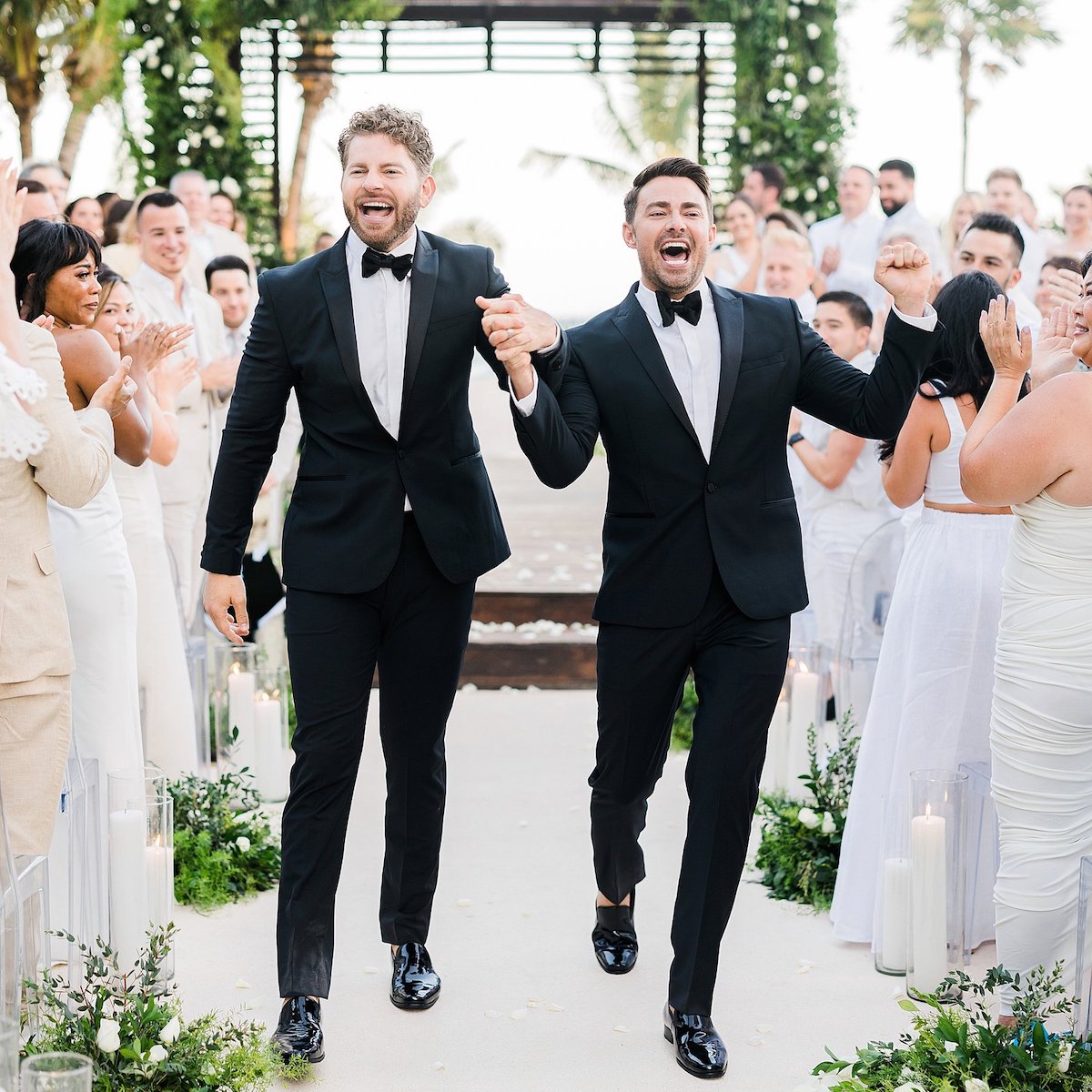 Two grooms in black tuxedos walk down the aisle hand-in-hand, surrounded by applauding guests in a stunning tropical outdoor setting, reminiscent of glamorous celebrity weddings.