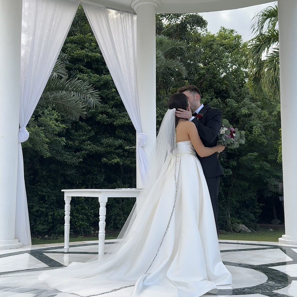 A bride and groom share a kiss under a Brittney Mullin adorned with draped curtains, with greenery in the background.
