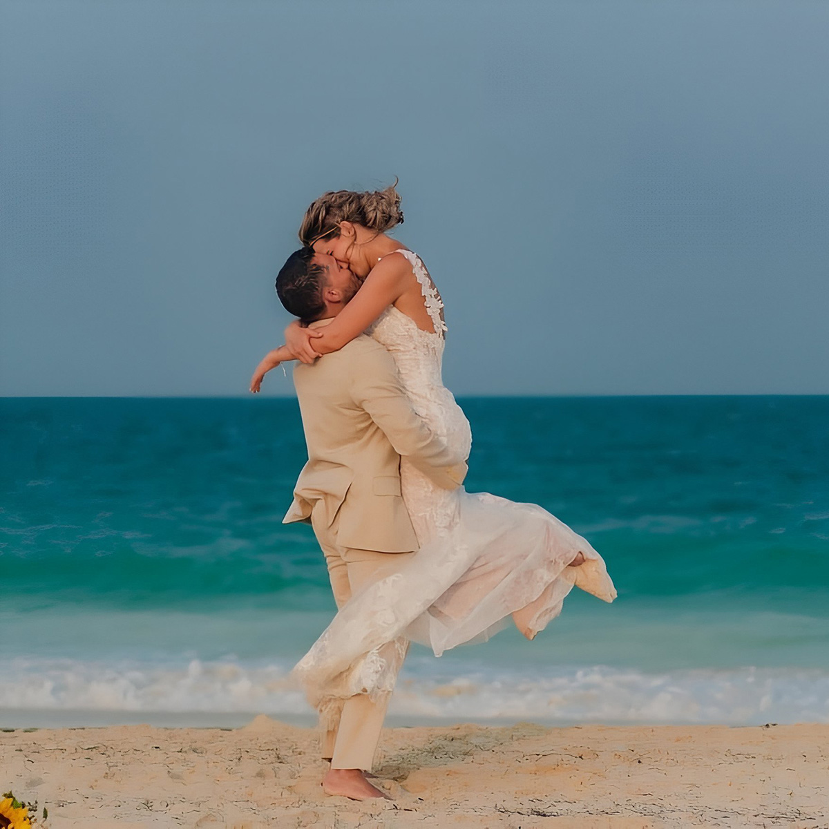 A newlywed couple in wedding attire, Candace and Sebastian, embrace joyfully on a sandy beach near the ocean. The groom, in a beige suit, lifts the bride, in a white dress, off the ground.