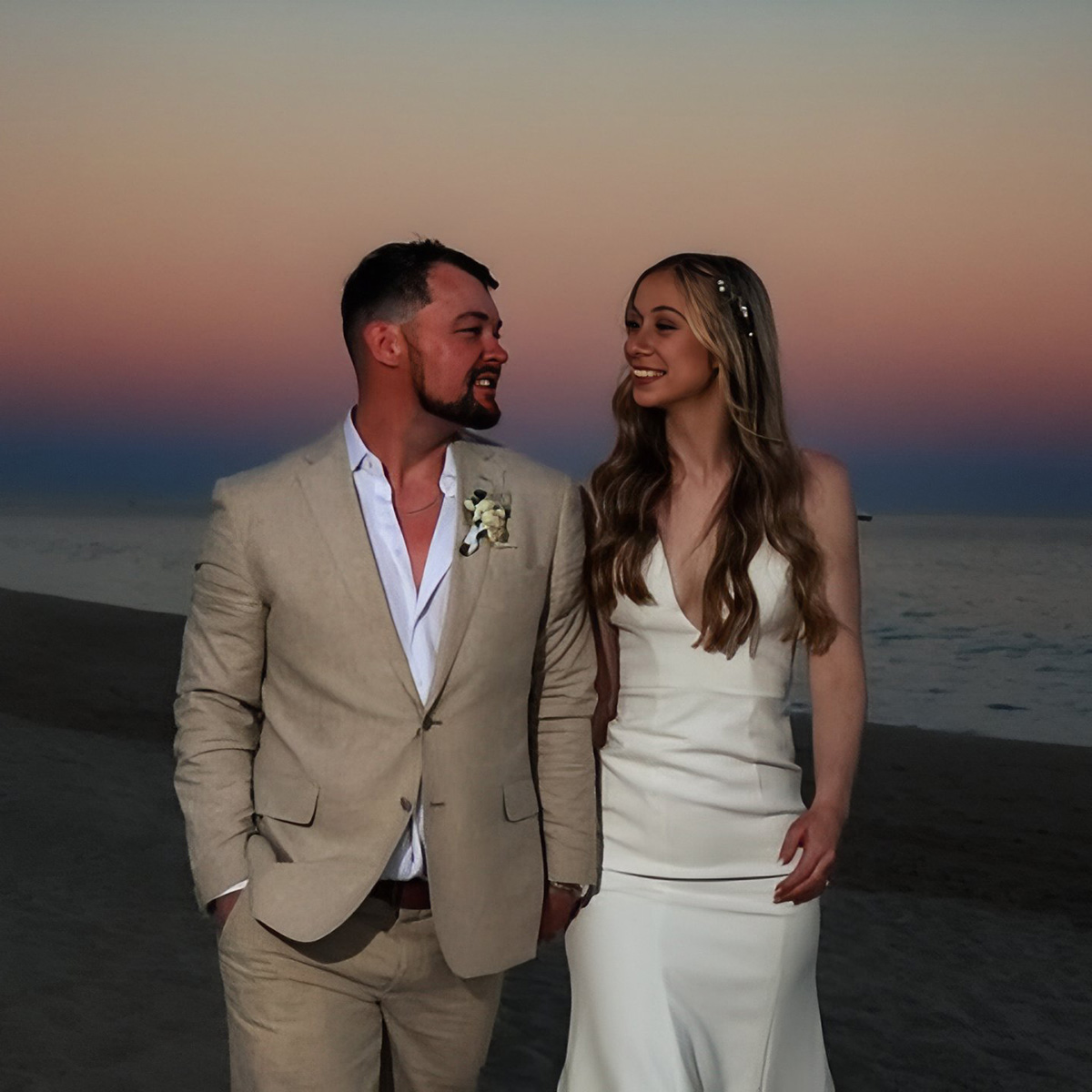 A man in a tan suit and a woman in a white dress walk hand in hand on a beach at sunset, against a backdrop of Emily and Dakota.
