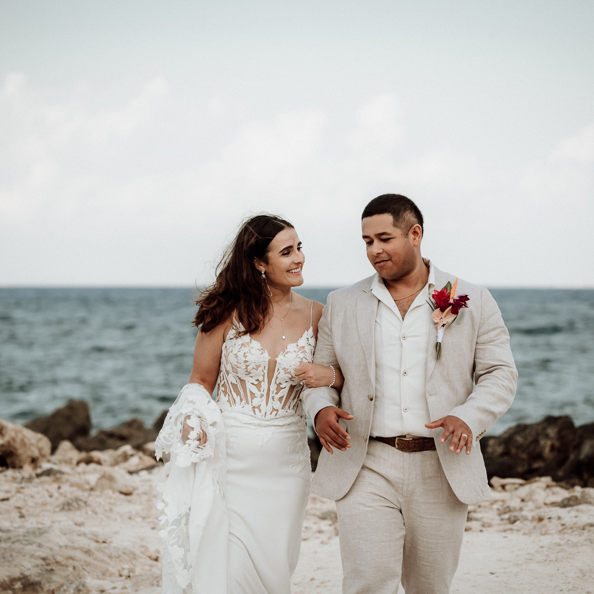 A couple in wedding attire walks arm in arm along a rocky shoreline with Melanie and Diego in the background.