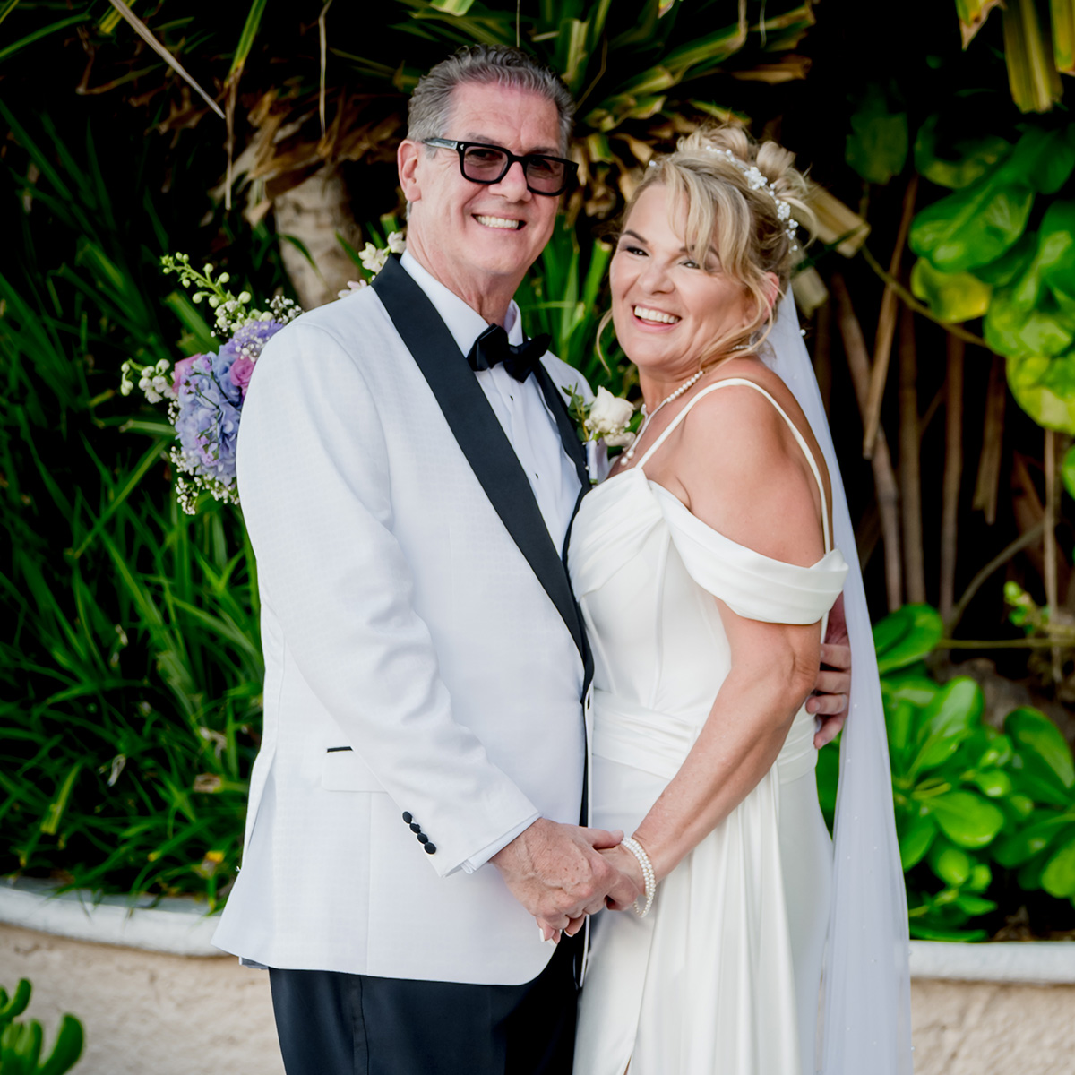 A couple dressed in formal attire, with the man in a white tuxedo and the woman in a white wedding dress, smile and hold hands in an outdoor setting with greenery in the background.