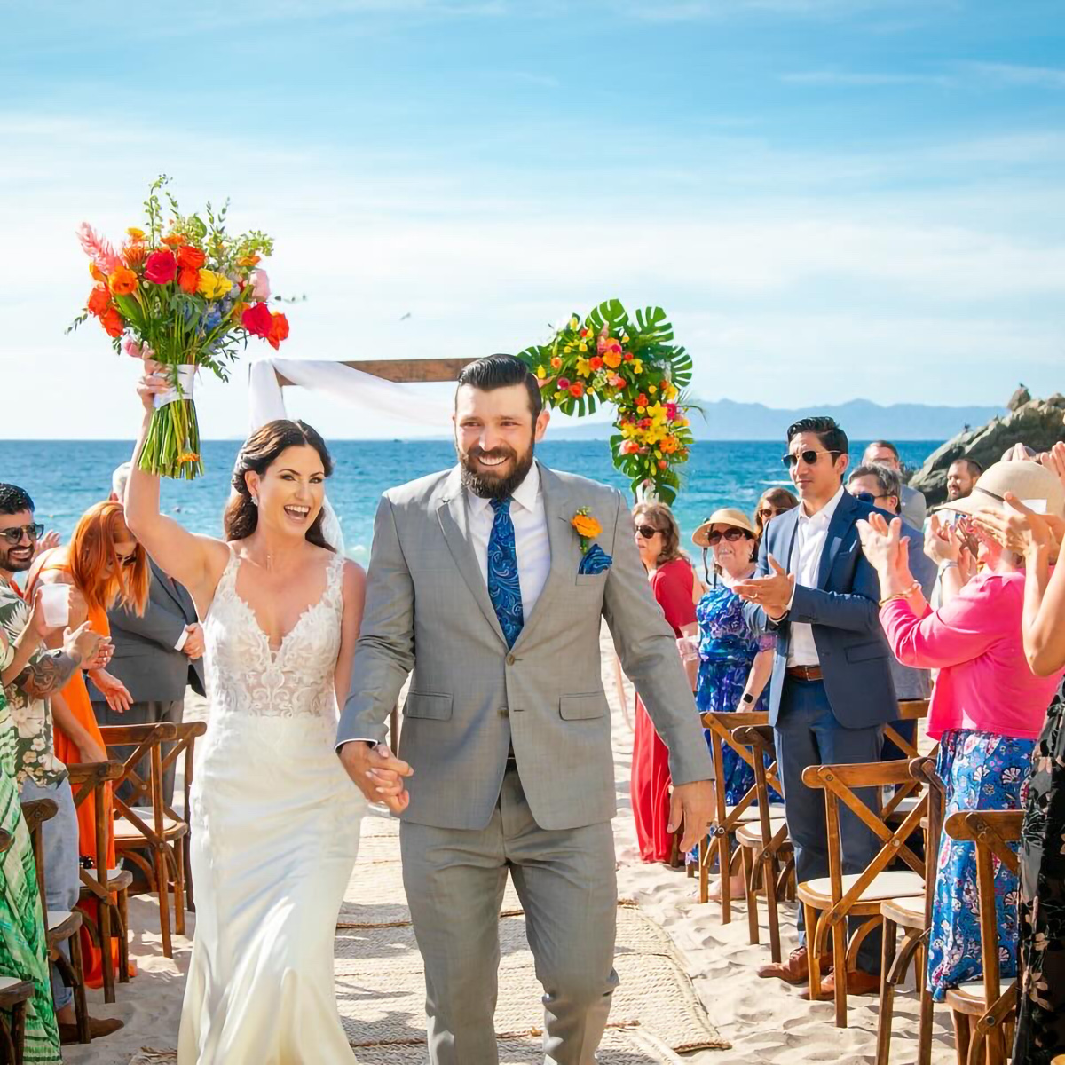 A newlywed couple, Molly and Zachary, walk down an aisle on a beach, holding hands. The bride raises her bouquet while guests clap and cheer. The ocean and blue sky serve as the backdrop.