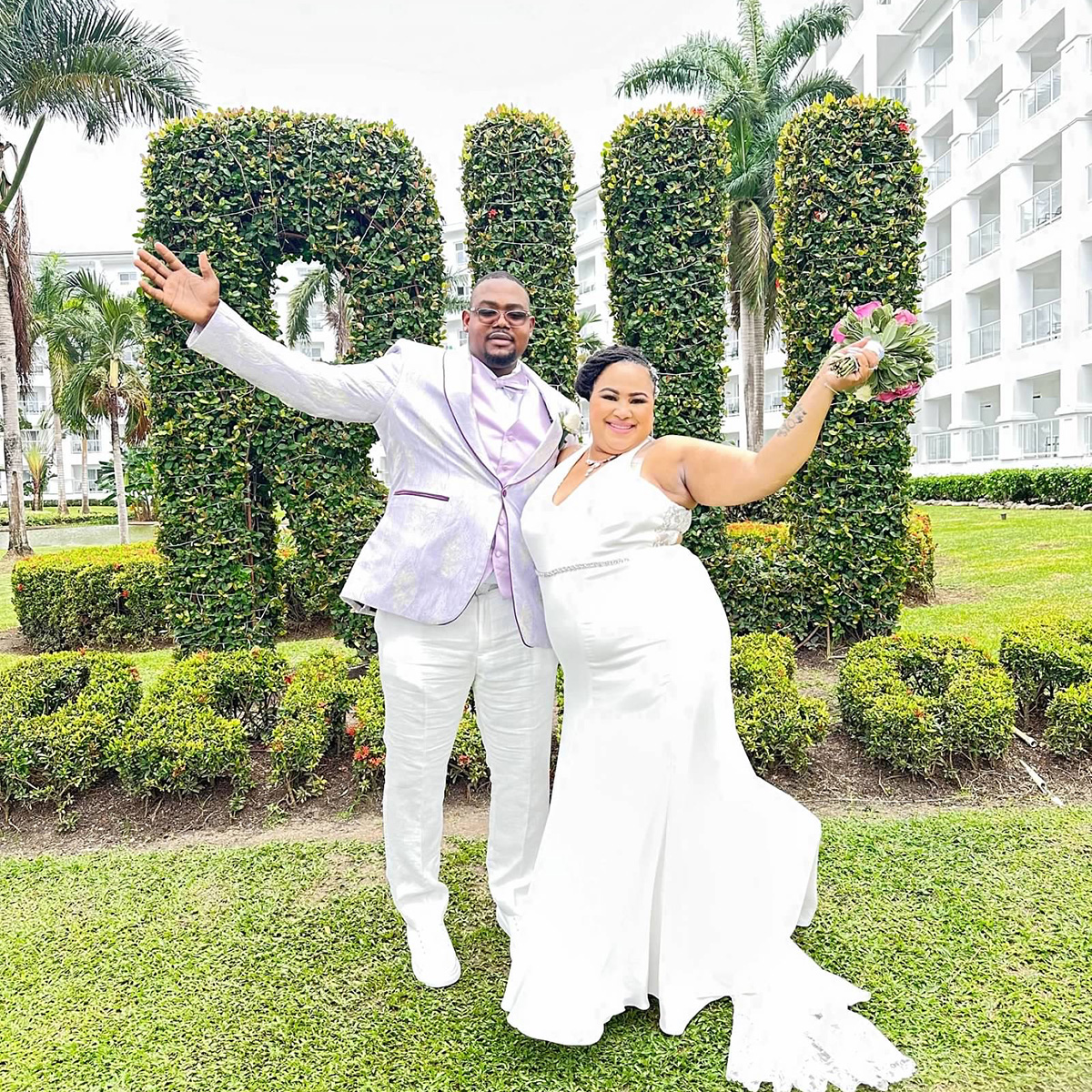 A newlywed couple poses outside in front of large greenery shaped into the word "Antoinette Coates." The groom wears a white suit with a light purple waistcoat, and the bride wears a white gown.