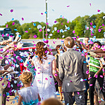 A couple in wedding attire walks through a crowd throwing colorful confetti outdoors.