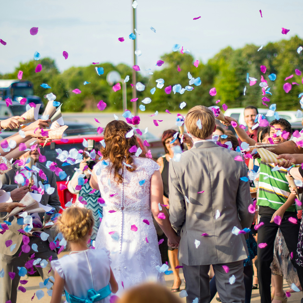 A couple in wedding attire walks through a crowd throwing colorful confetti outdoors.