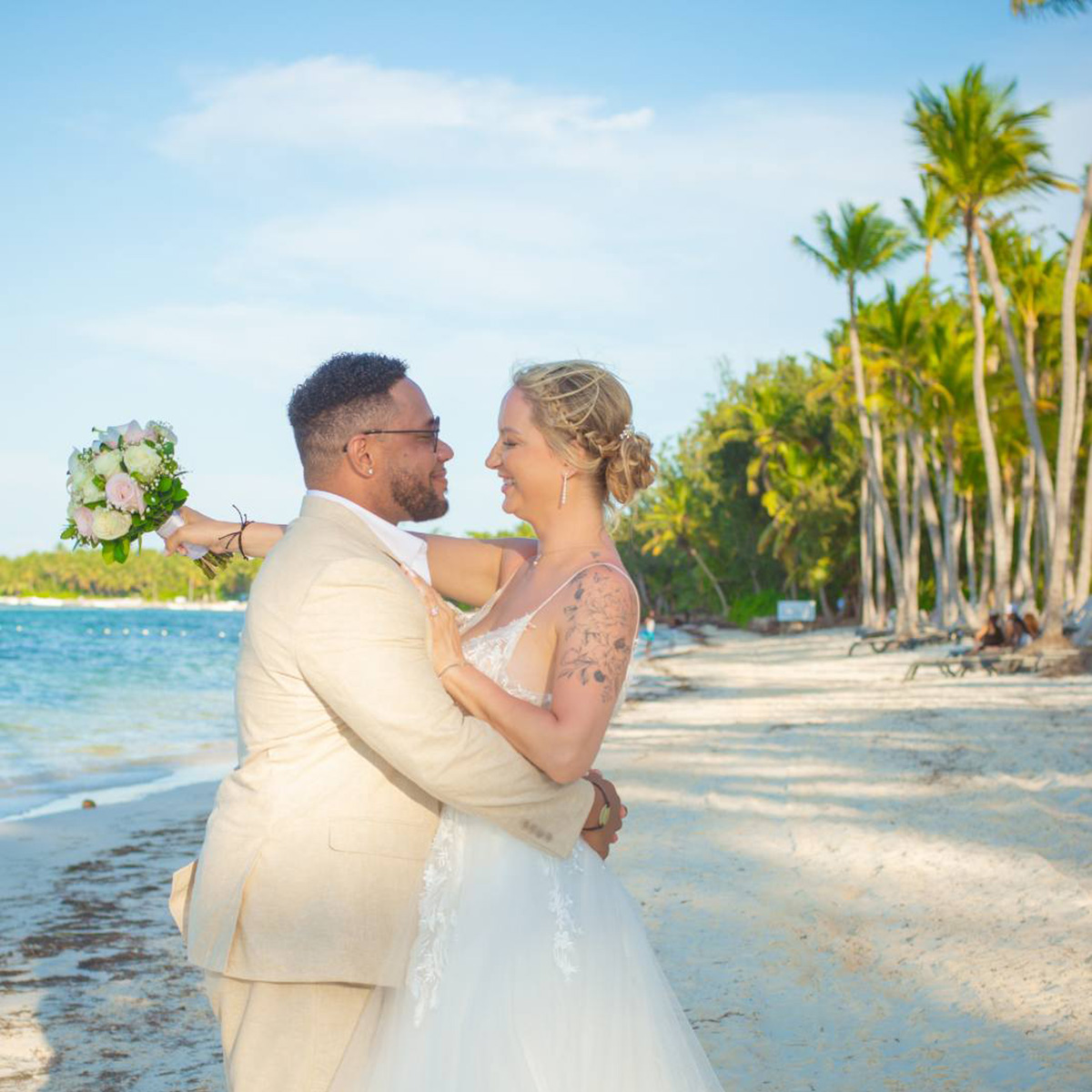A couple in wedding attire embraces on a tropical beach. The groom is in a beige suit, and the bride holds a bouquet, both smiling with a backdrop of palm trees and ocean.