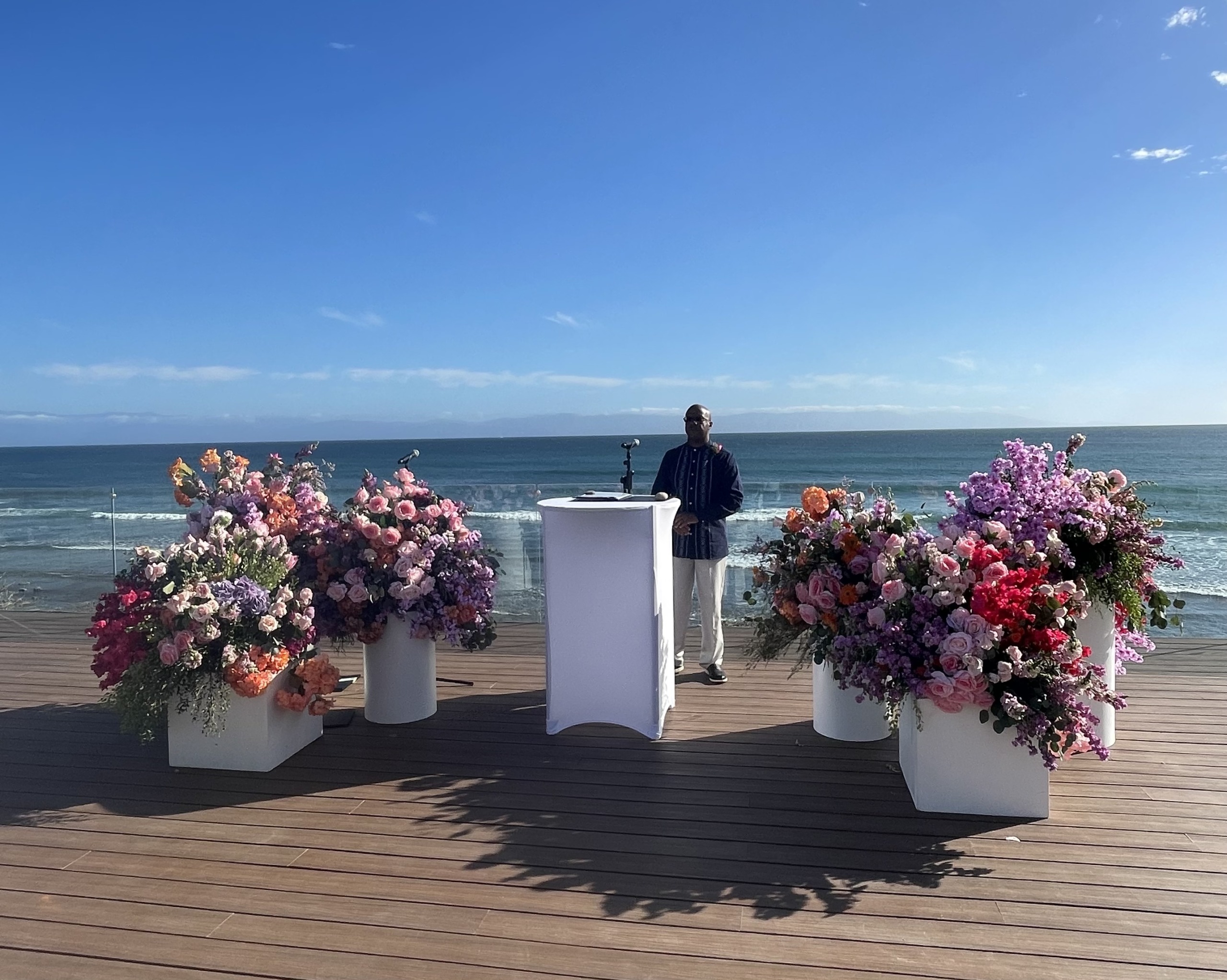 A person stands behind a podium on a deck overlooking the ocean, surrounded by colorful flower arrangements on a sunny day.