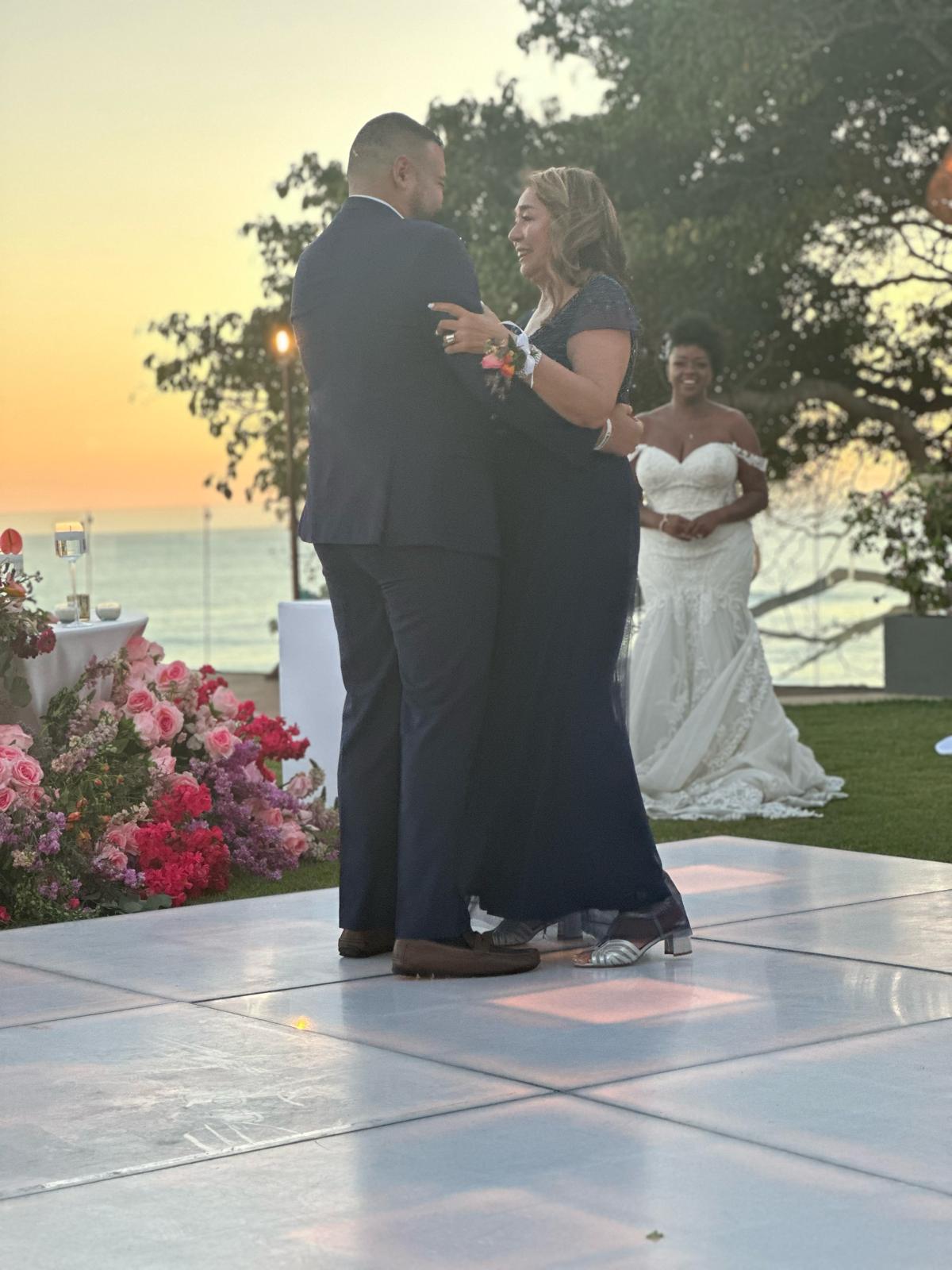 A couple dances together on a white floor at an outdoor event during sunset, while a bride in a white gown stands in the background, smiling. Flowers and a drink are arranged on a nearby table.