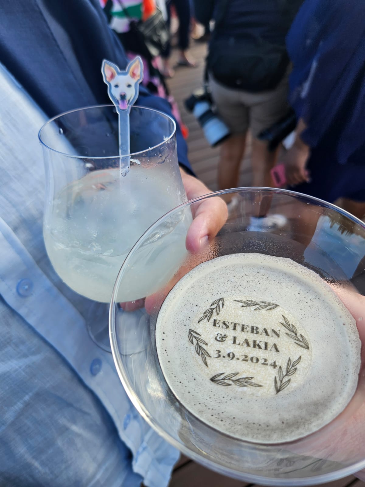 Person holding a cocktail with a dog stirrer and a drink featuring a foam decoration with the text "Esteban & Lakia 3-9-2024" at a crowded event.