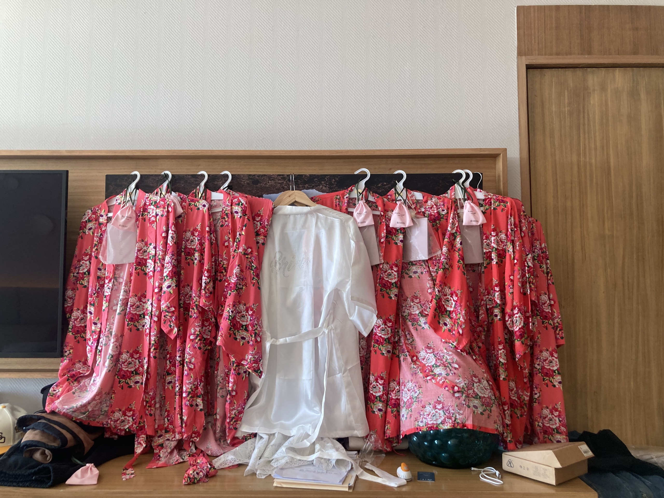 A row of six pink floral robes and one white robe hang on a rack. Various items are scattered on the table below, including boxes and clothes.