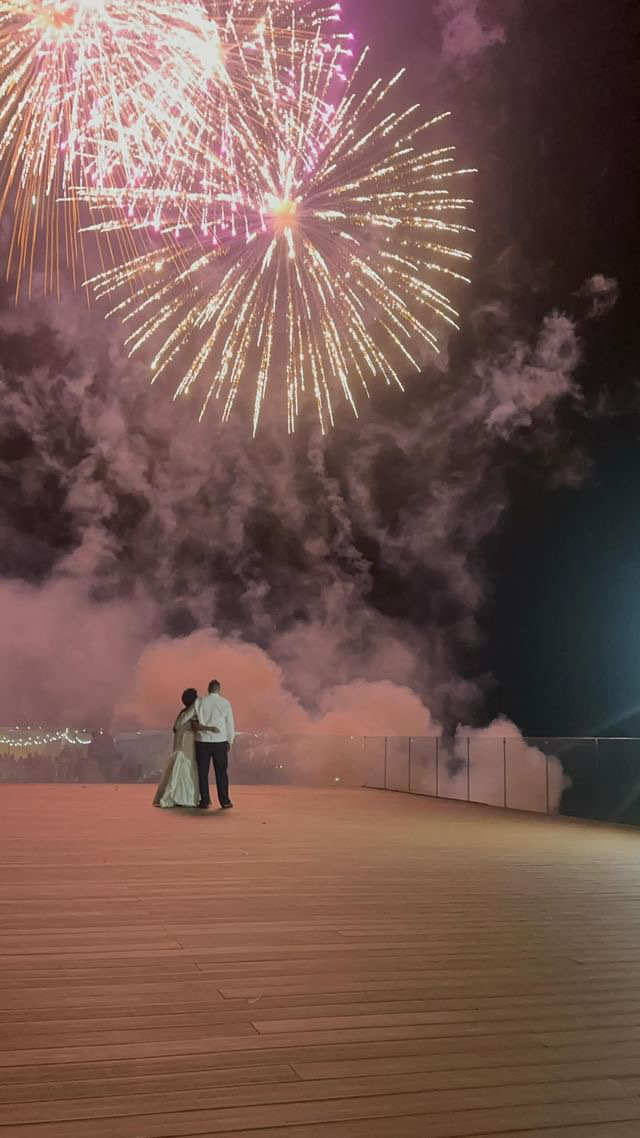 A couple in formal attire watches fireworks on a wooden deck at night, with colorful explosions and smoke lighting up the sky.