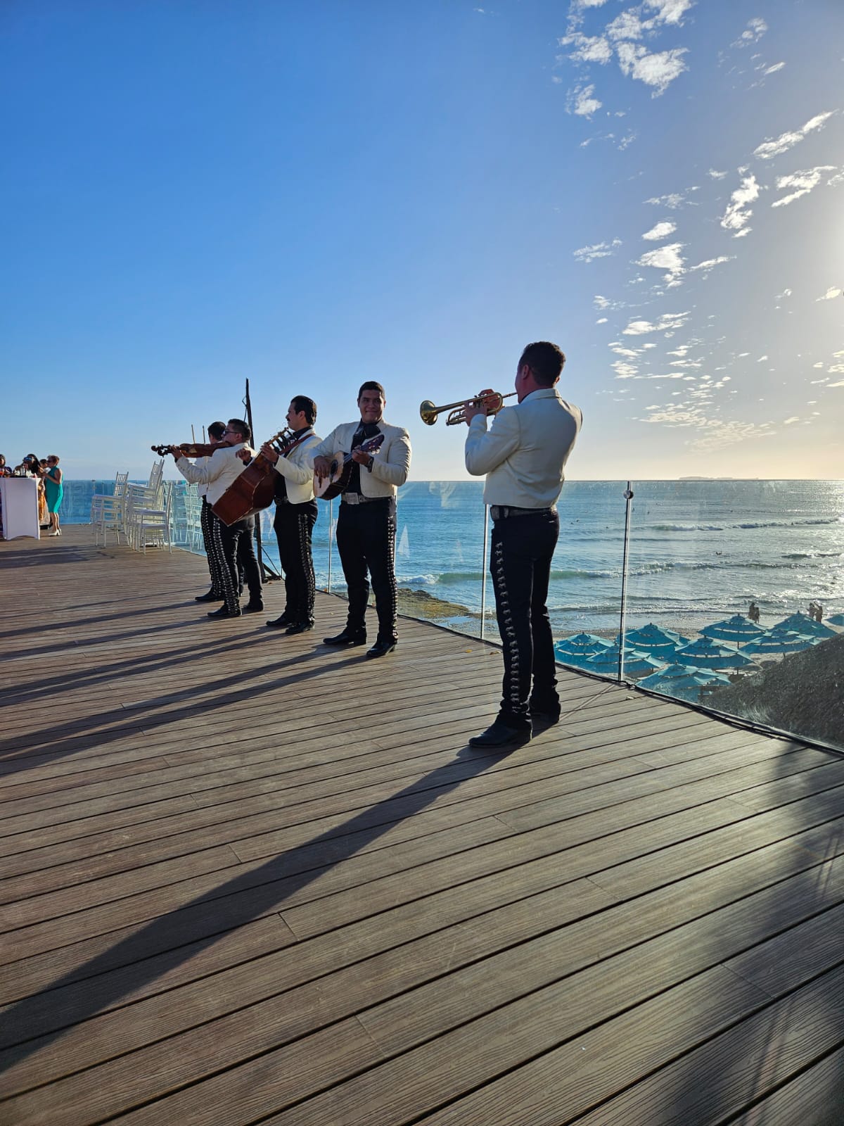 A mariachi band performs on a wooden deck overlooking the ocean under a bright, clear sky.