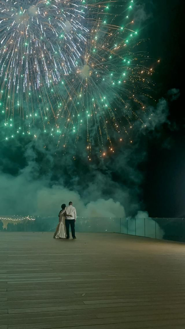 A couple stands on a wooden platform at night, watching a large display of green and white fireworks in the sky.