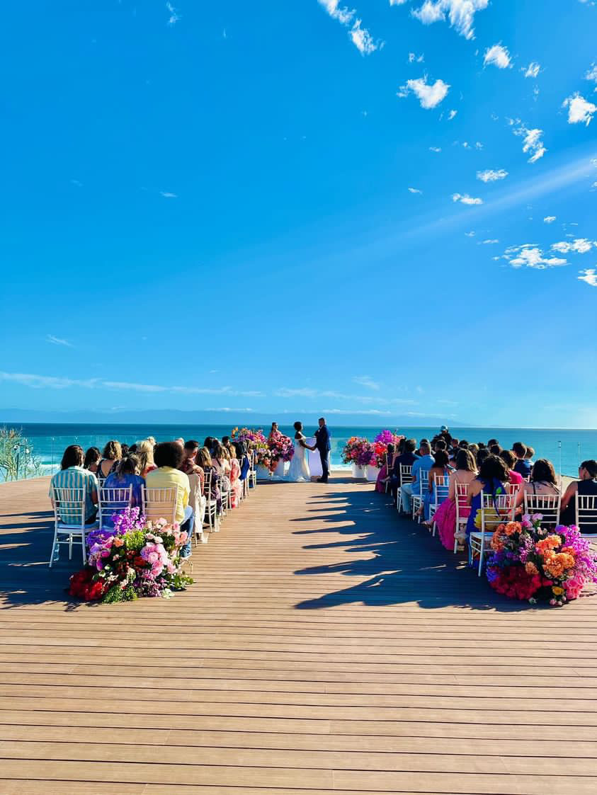 A wedding ceremony on a wooden deck by the ocean, with an aisle lined with colorful flowers and guests seated on either side under a clear blue sky.