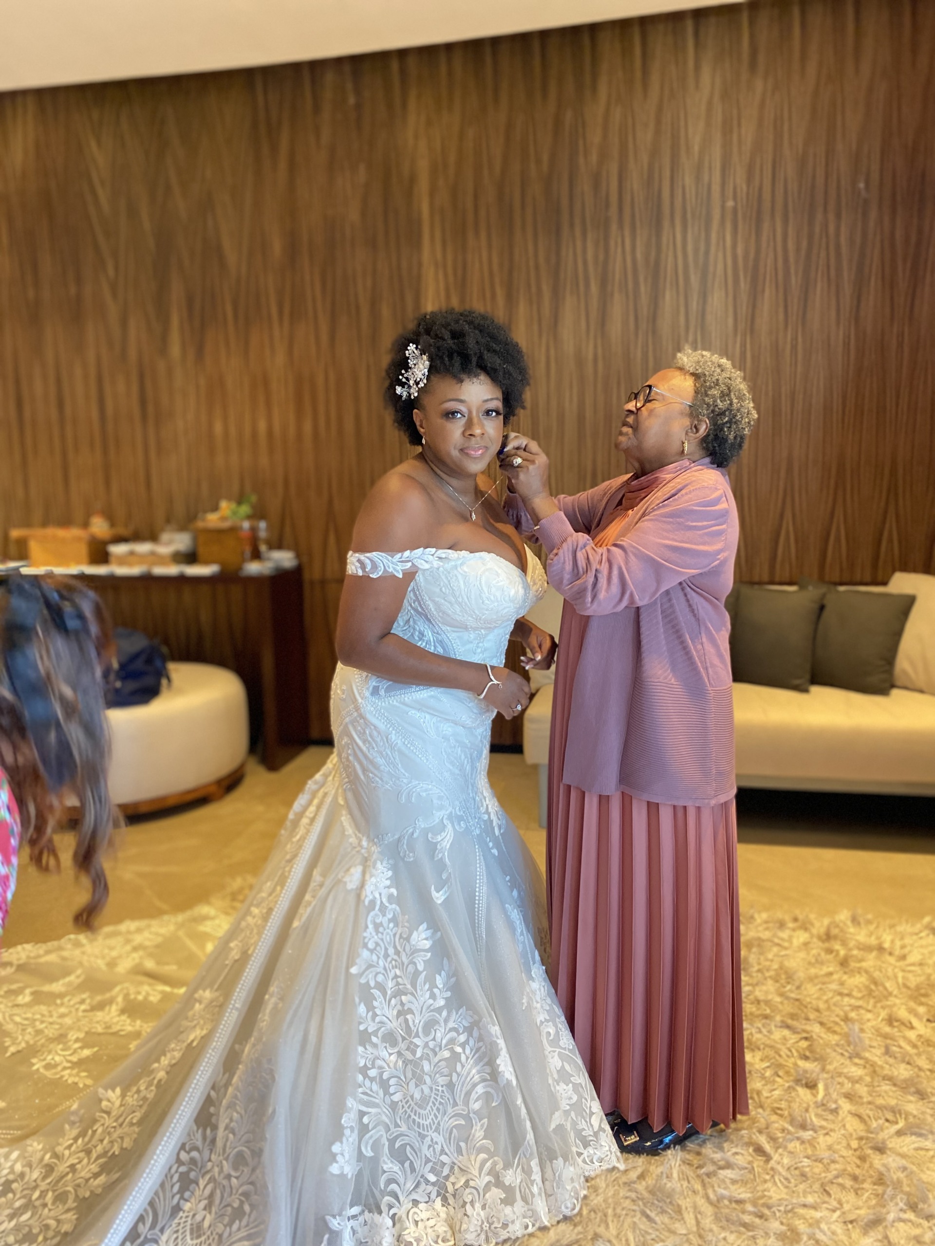 A woman in a wedding dress stands while another woman adjusts her necklace. They are in a room with wooden walls and a sofa in the background.