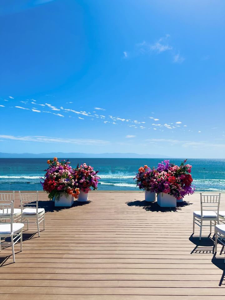 A wooden deck overlooking the ocean, adorned with two large floral arrangements on either side, and white chairs set for an event under a clear blue sky.