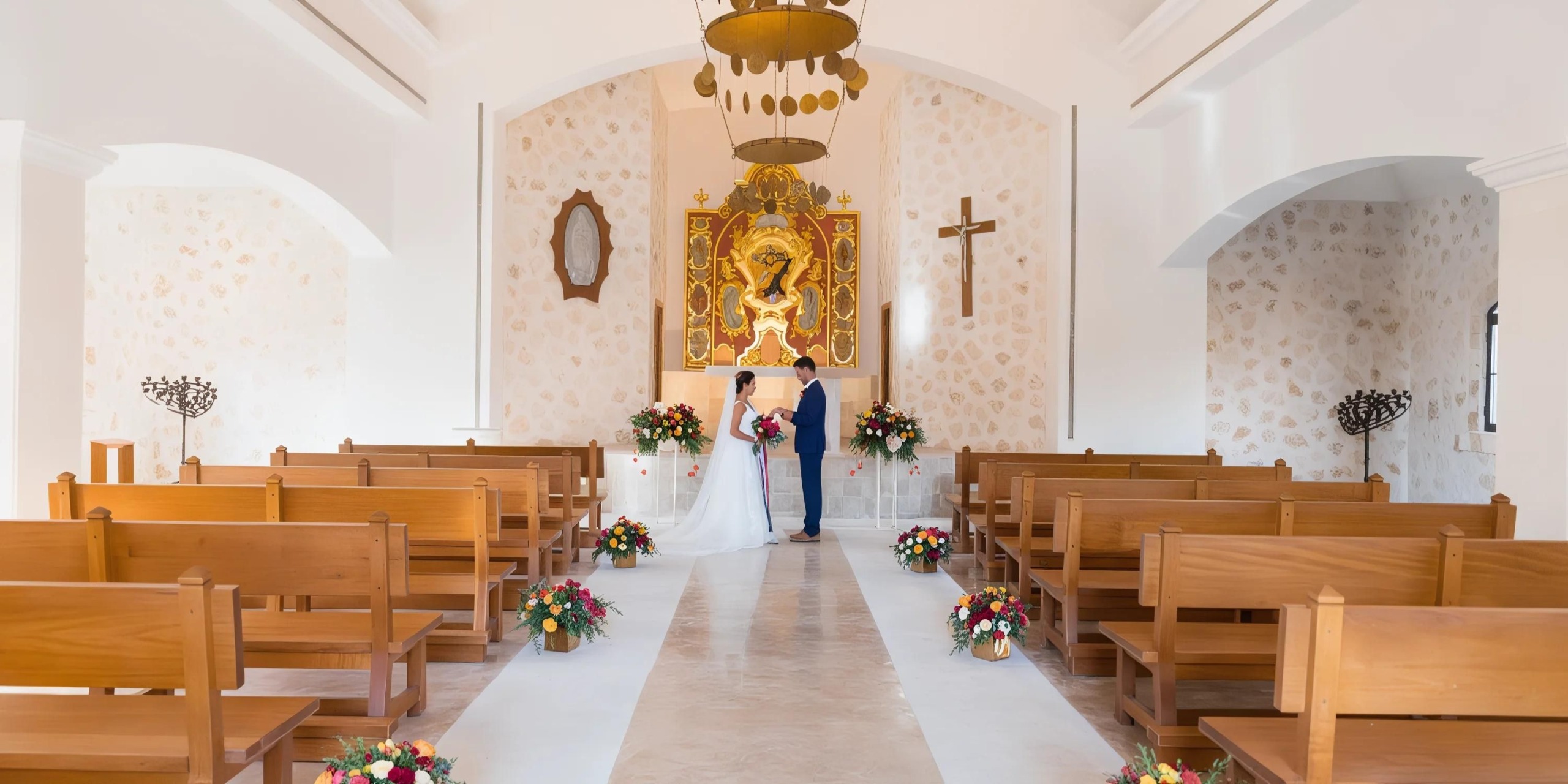 A bride and groom stand at the altar in a small chapel decorated with floral arrangements and a large religious painting, facing each other and holding hands.