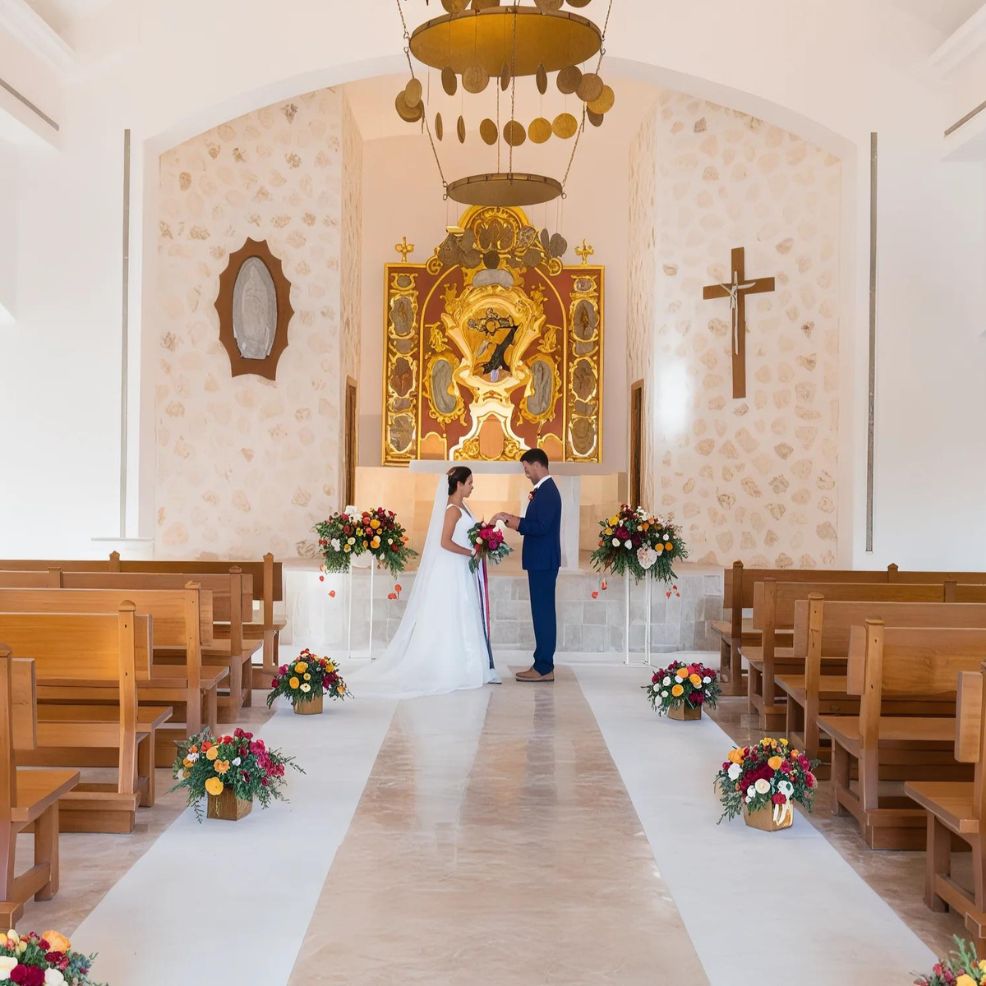A bride and groom stand at the altar in a small chapel, holding hands and facing each other. The chapel is decorated with floral arrangements and features a crucifix and religious artwork.