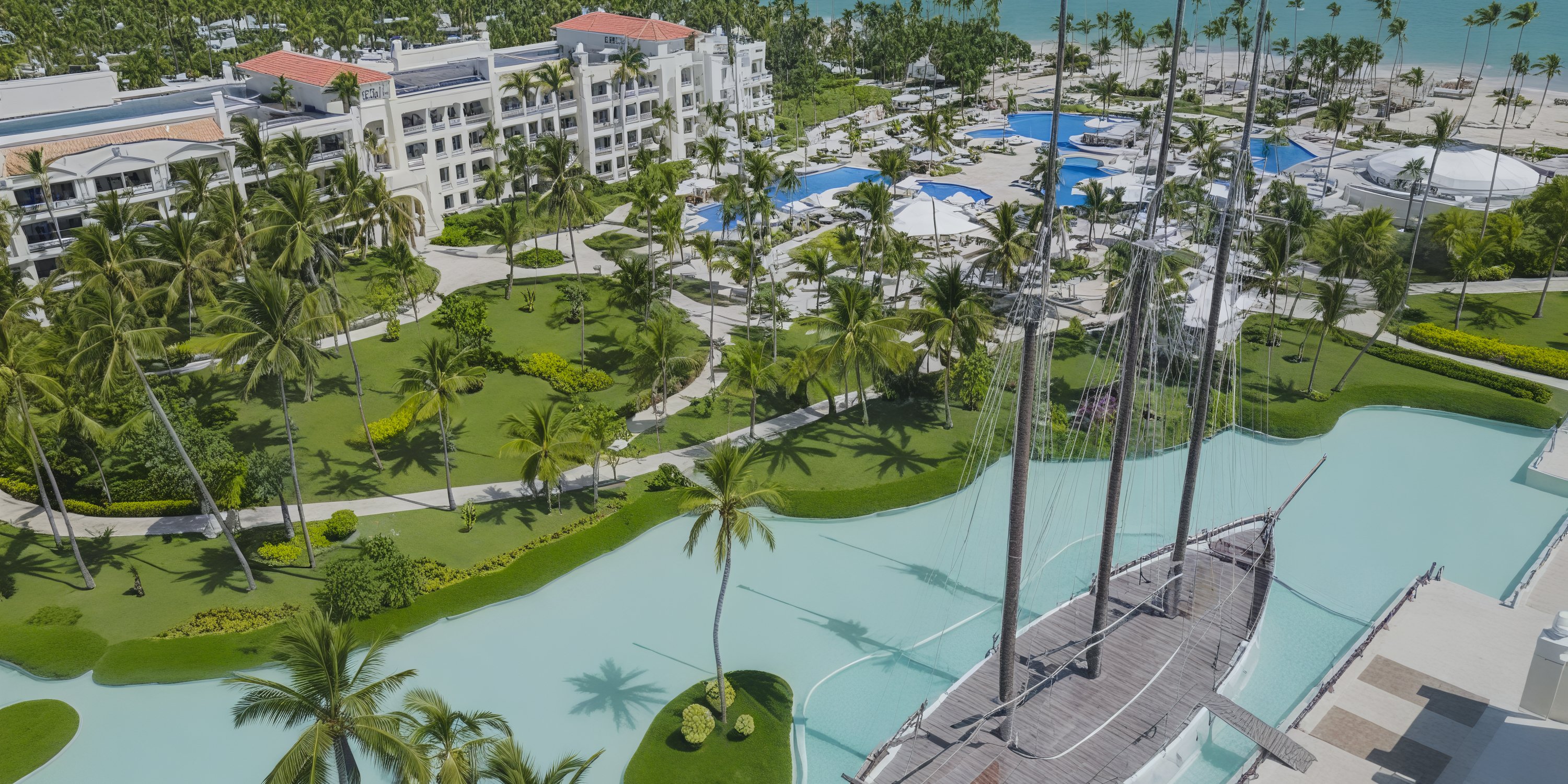 A breathtaking aerial view of JOIA Bavaro at the Iberostar resort, featuring stunning pools, lush gardens, and a charming boat in a turquoise canal—an ideal setting for an unforgettable destination wedding.