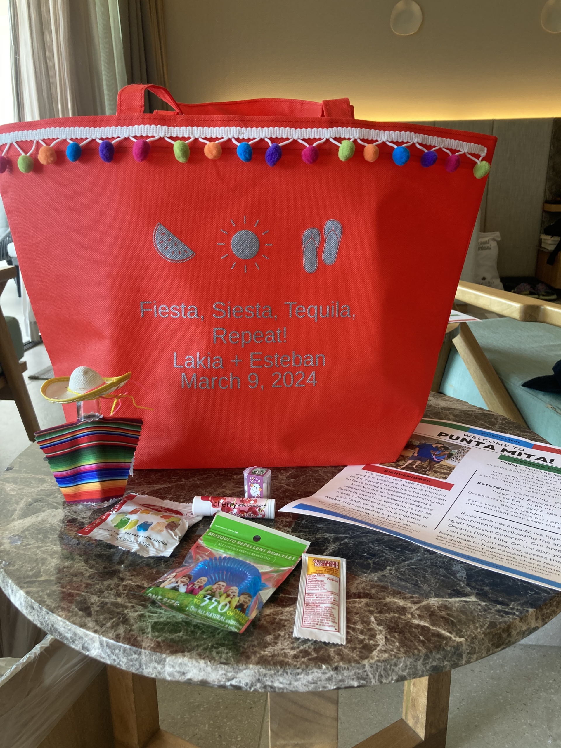 A gift bag with a festive design and the text "Fiesta, Siesta, Tequila, Repeat! Lakia + Esteban March 9, 2024" contains various items, including snacks and a pamphlet, on a marble surface.
.