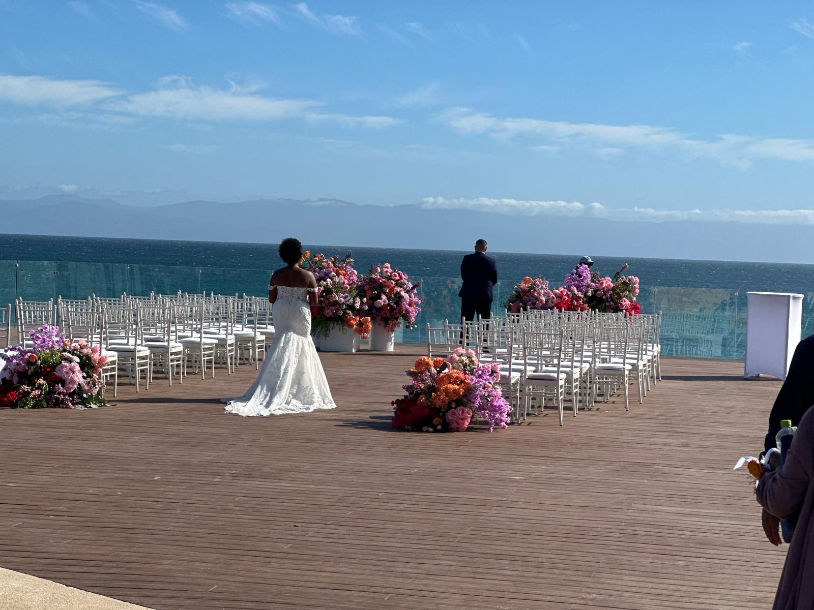 A bride in a white dress stands on a wooden platform decorated with flowers and white chairs, overlooking the sea, with a man in a suit in the background.