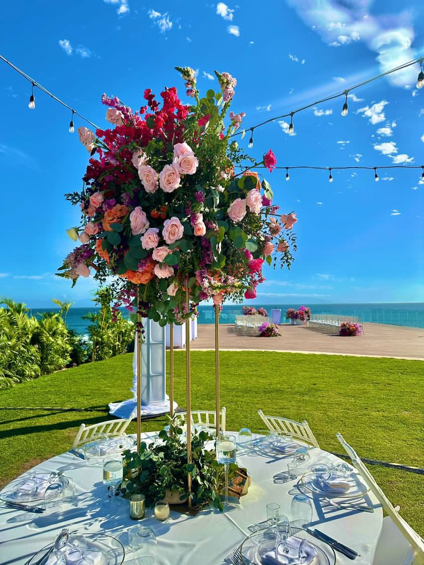A decorative table centerpiece with pink, red, and white flowers under string lights on a lawn overlooking the ocean.