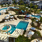 An aerial view of a resort pool area featuring winding pools, sun loungers with umbrellas, a small waterslide, and manicured landscaping. Several buildings surround the area, with palm trees scattered throughout.