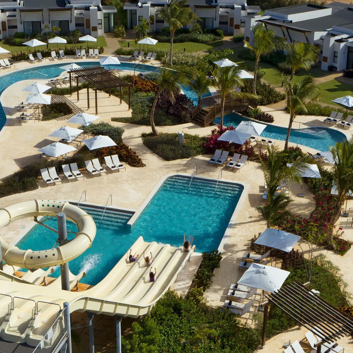 An aerial view of a resort pool area featuring winding pools, sun loungers with umbrellas, a small waterslide, and manicured landscaping. Several buildings surround the area, with palm trees scattered throughout.