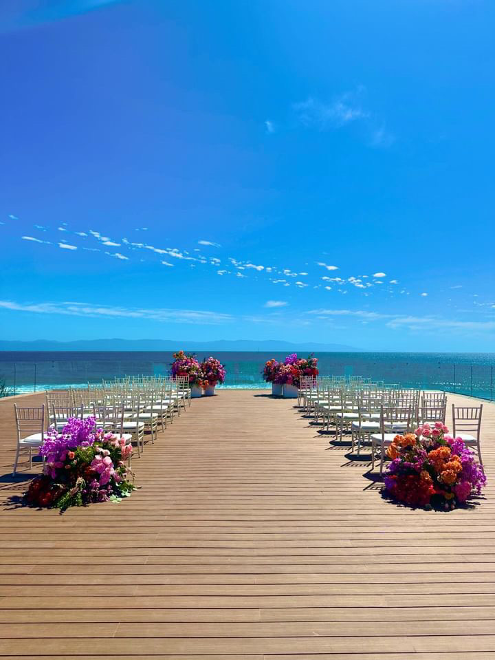 Outdoor wedding ceremony setup with rows of white chairs facing an ocean view. Colorful floral arrangements line the wooden aisle, and clear blue sky is above.