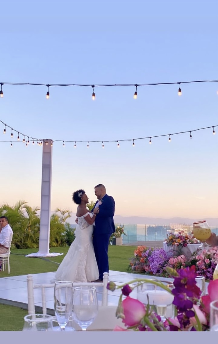 A bride and groom are dancing under string lights at an outdoor wedding reception with a colorful floral arrangement and a scenic ocean view in the background.