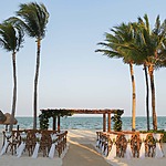 A beachside wedding setup in the all-inclusive Riviera Maya with a wooden altar, decorated chairs, and palm trees, overlooking the ocean under a clear sky.