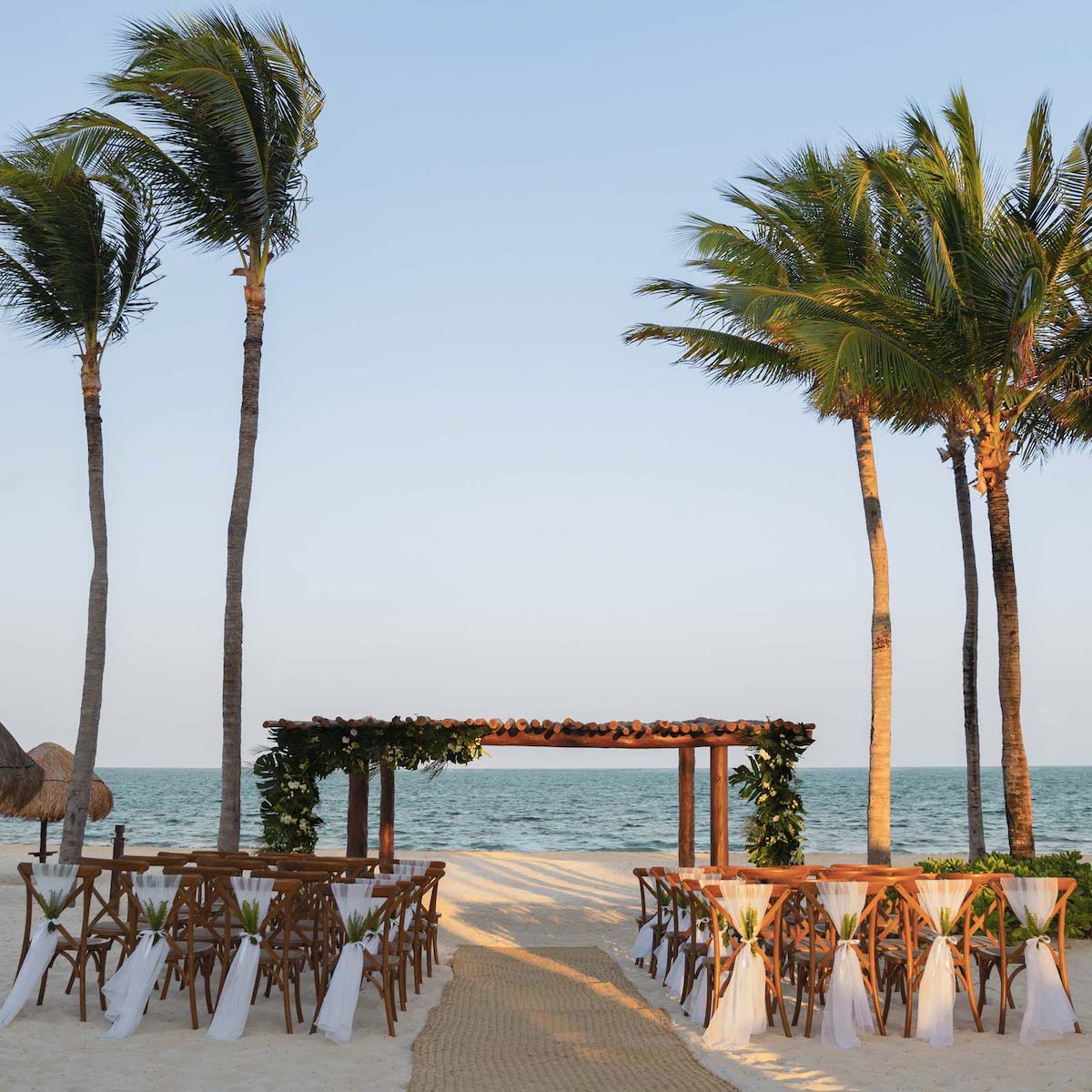 A beachside wedding setup in the all-inclusive Riviera Maya with a wooden altar, decorated chairs, and palm trees, overlooking the ocean under a clear sky.