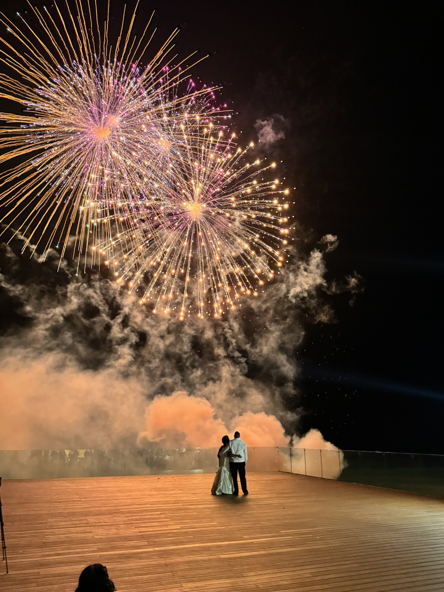 A couple stands close together on a boardwalk at night, watching a vibrant fireworks display light up the sky. Smoke and sparks from the fireworks add a dramatic effect.