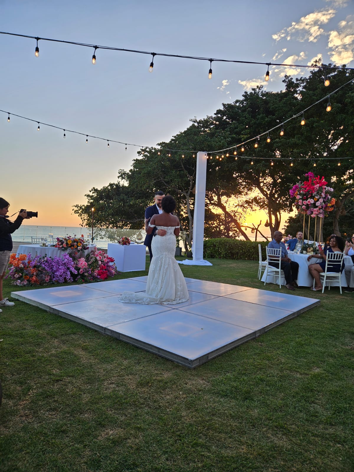 A bride and groom share their first dance on an outdoor dance floor surrounded by floral decorations and string lights at sunset, while a photographer captures the moment.