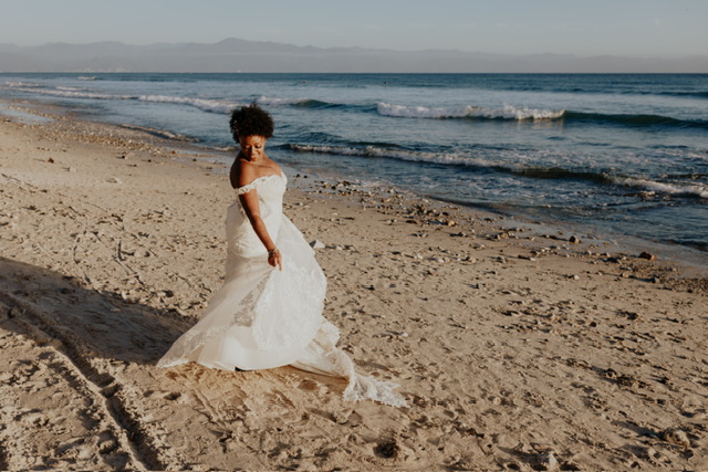 A person in a white off-the-shoulder wedding dress stands on a sandy beach with waves crashing in the background.