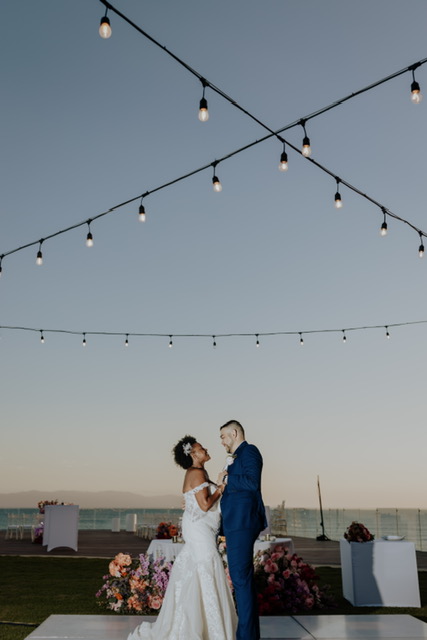A couple in formal attire shares a dance under string lights on an outdoor platform decorated with flowers, with a clear sky in the background.