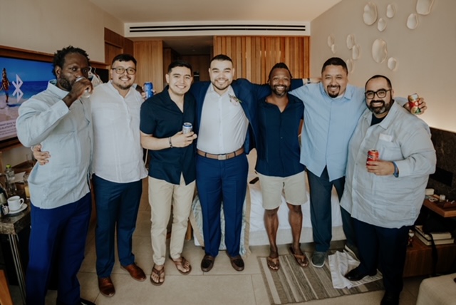 A group of seven men standing together in a room, smiling at the camera. Some hold beverages. They are wearing casual and semi-formal clothing. The room has wooden paneling and wall decorations.