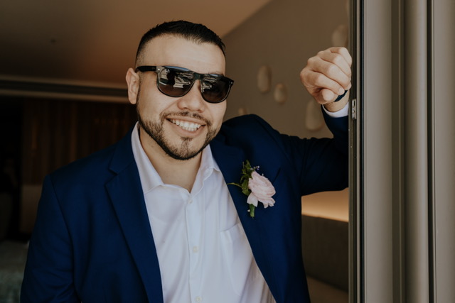 A man wearing sunglasses and a blue suit, with a white shirt and a pink boutonniere, leans against a door frame, smiling.