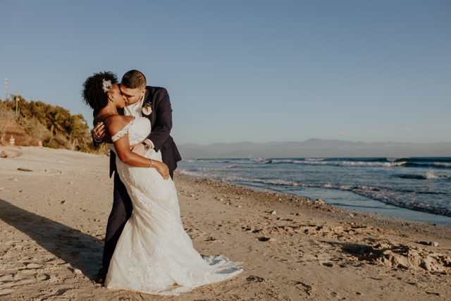 A couple in wedding attire shares a kiss on a sandy beach with the ocean and distant mountains in the background.