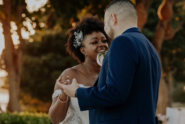 A couple, both dressed formally in a wedding dress and suit, share a dance outdoors with trees blurred in the background.
