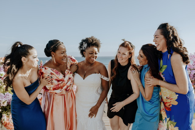 A bride in a white dress and five women in colorful dresses stand outdoors, laughing and smiling together.