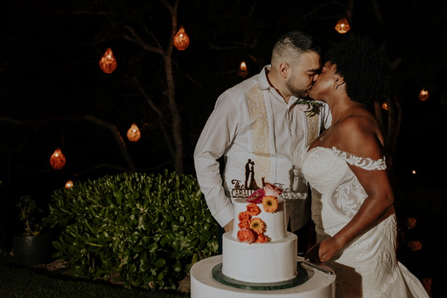 A bride and groom kiss next to a wedding cake decorated with flowers and a silhouette cake topper. The background features hanging lights and a garden setting at night.