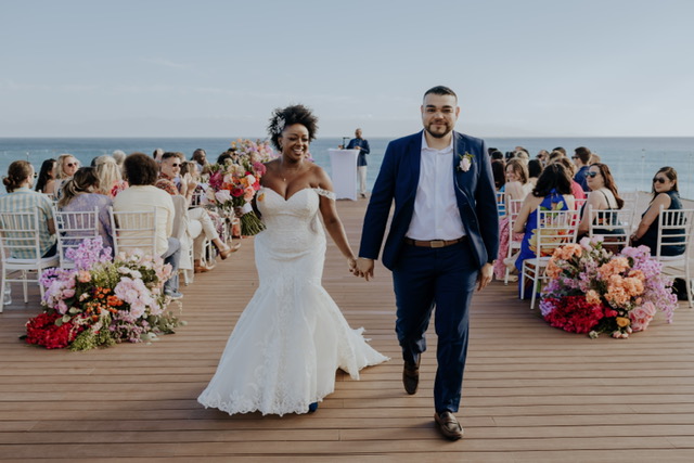 A bride and groom hold hands and smile as they walk down the aisle at an outdoor wedding ceremony. Guests are seated on either side of the aisle, with floral decorations on the ground.
