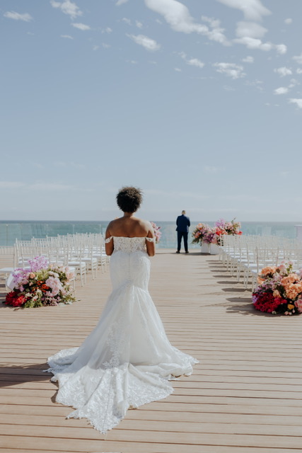 A bride in a white gown walks down an outdoor aisle towards a groom standing at the end. The aisle is decorated with flowers, and white chairs are set on either side under a clear sky.